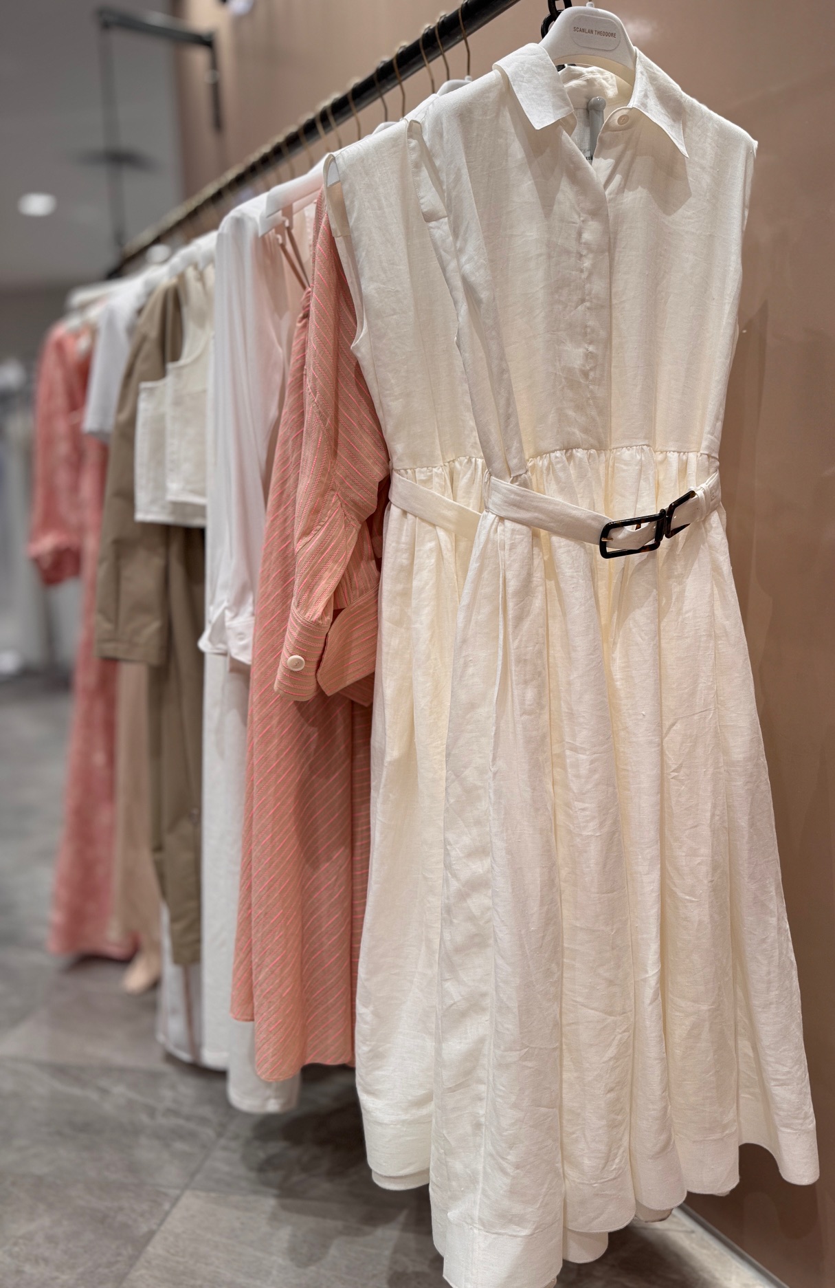 A row of white and blush linen dresses hanging neatly in a boutique, soft daylight revealing their natural weave.