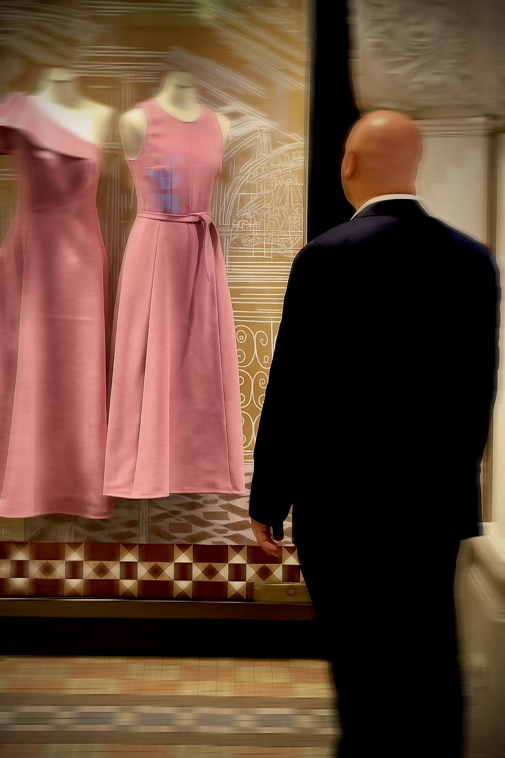 White collared man in suit staring at two pink dresses in store window.
