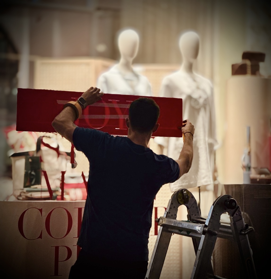 Worker installing red store sign in front of fashion mannequins.