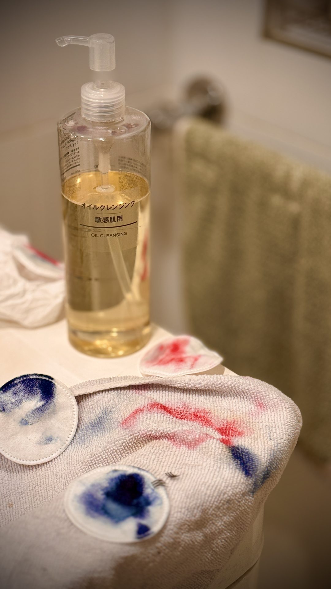 Oil cleanser bottle with makeup-stained cotton pads and towel on a bathroom counter.