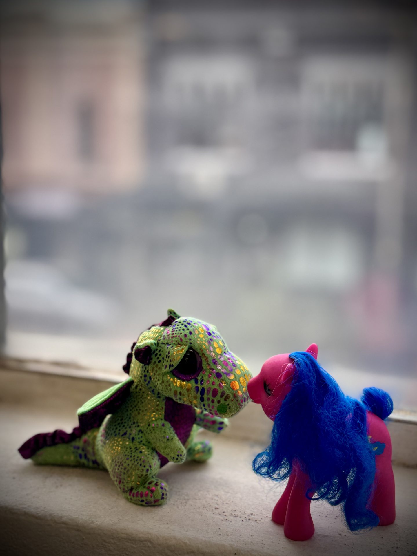 A green plush dragon touching noses with a pink toy pony on a window ledge.