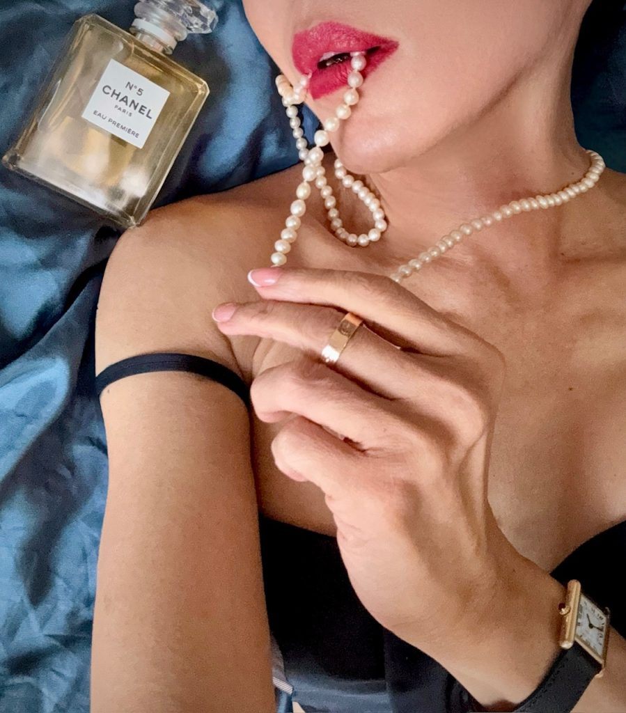 Close-up of red lips holding a pearl necklace beside a Chanel No.5 Eau Première bottle.