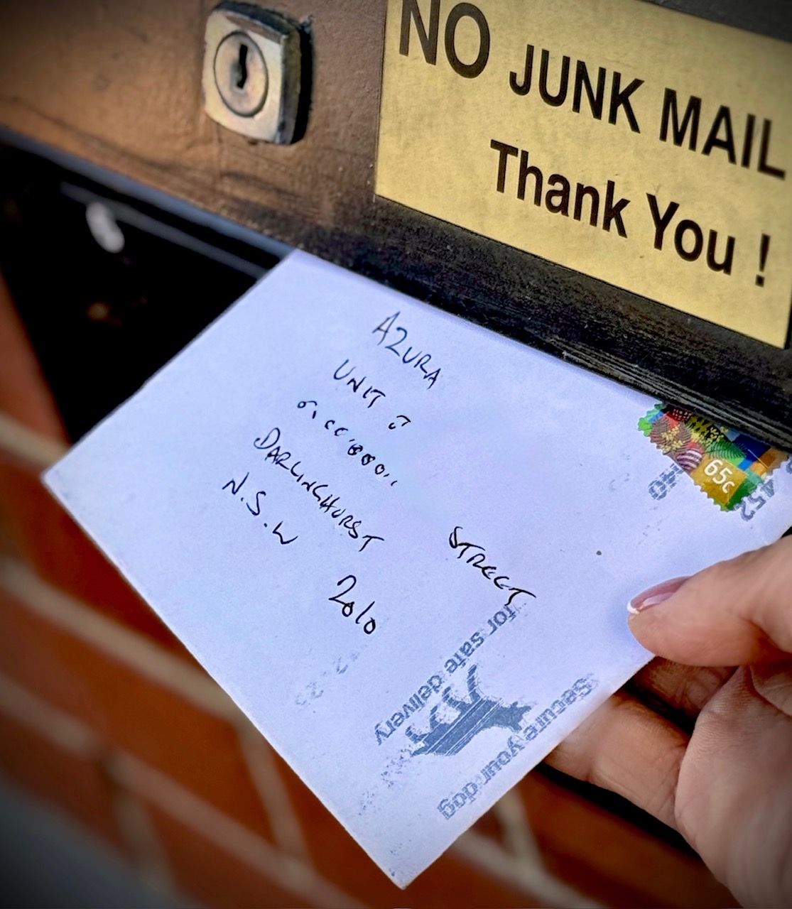 Hand pulling a handwritten envelope from a mailbox beneath a “NO JUNK MAIL” sign, with an Australian stamp and postmark.