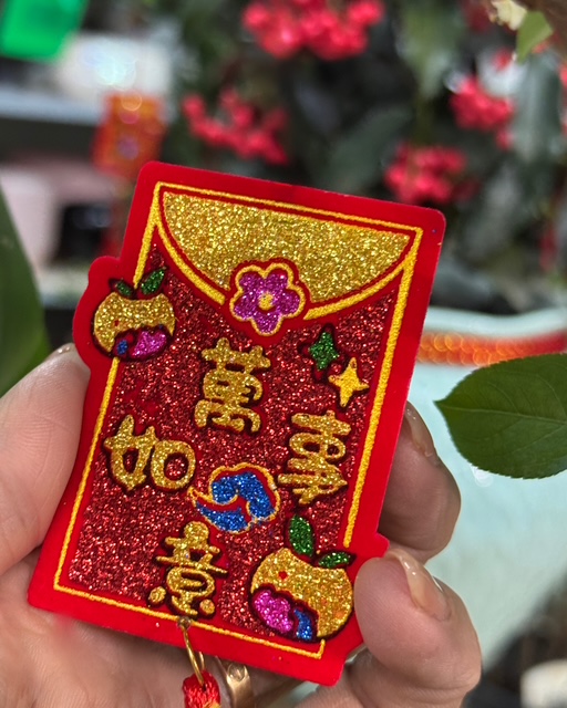 Glittering red Lunar New Year angpao ornament held between fingers with gold Chinese characters.