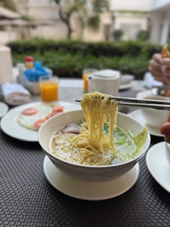 Chopstick lifting yellow egg noodles from a bowl of Thai noodle soup at an outdoor breakfast table.